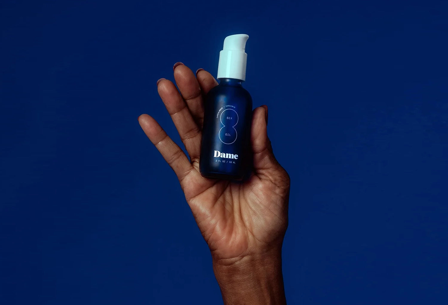 Óleo de massagem Dame Sex Oil num frasco azul — segurado na mão com fundo forte azul