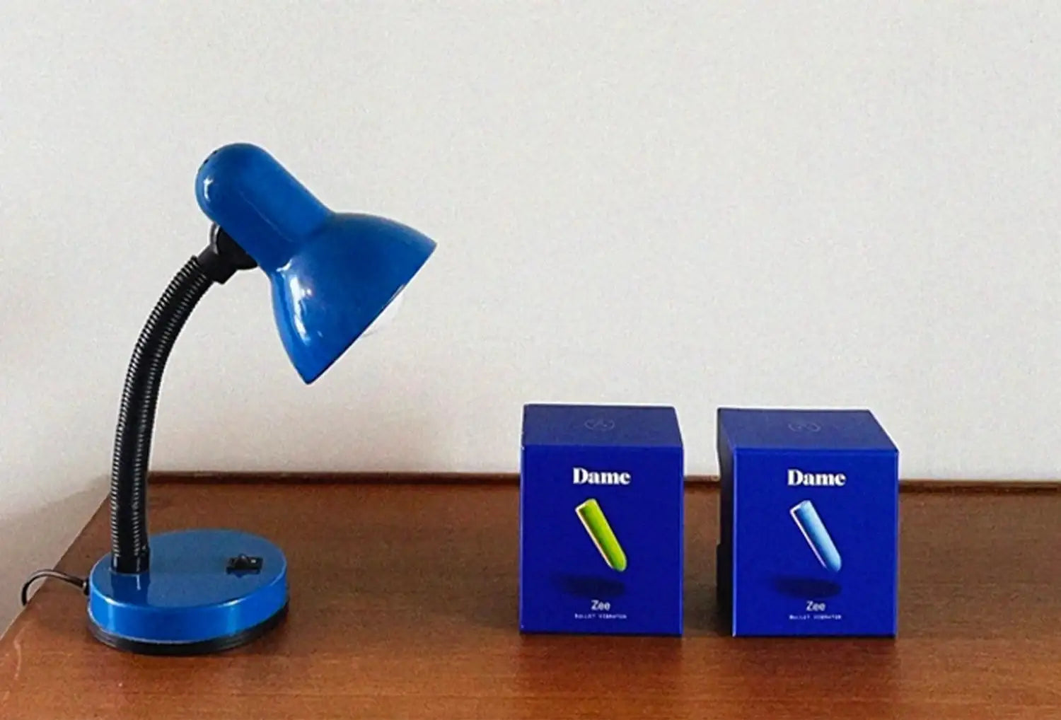 Vibrador bullet clitoriano Dame Zee em ABS nas cores pervinca e cítrico — duas embalagens azuis em cima de uma mesa de madeira escura ao lado de um canteiro azul