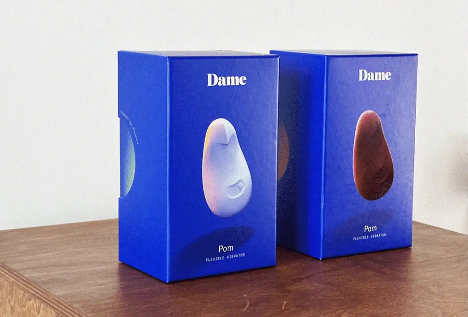Vibrador clitoriano Dame Pom em silicone de cores lavanda e ameixa — duas embalagens em cima de uma mesa de madeira escura