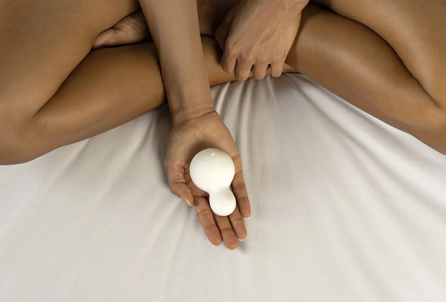 Vibrador clitoriano Iroha Yuki em silicone de cor branco nas mãos em zoom de uma mulher sentada em flor de lótus em cima de uma cama de algodão branco