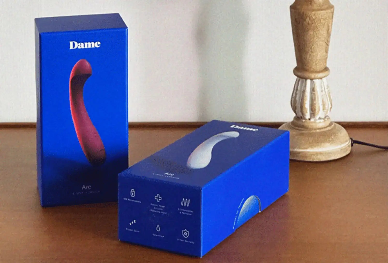 Vibrador de ponto g Dame Arc em silicone de cores lavanda e ameixa — duas embalagens azuis em cima de uma mesa de madeira escura.